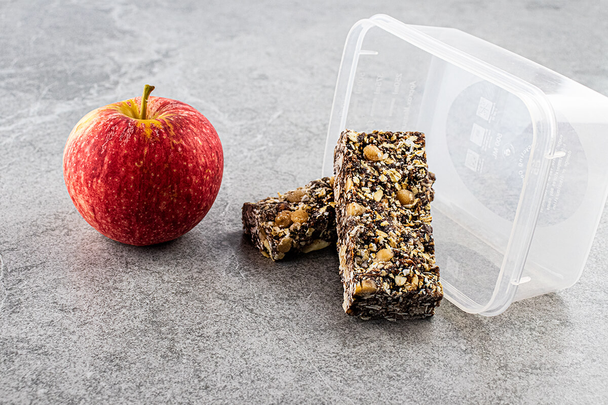 Grey slate background with lunchbox inspired setting (apple and reusable container used as props) with Choc Almond Fudge Museli Bar